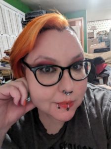 Tart, a pale femme with short neon orange hair, facing the camera with heavy makeup on